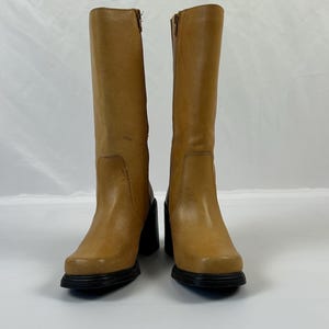 Puede incluir: Un par de botas altas de cuero color canela con puntera cuadrada y tacones de bloque negros. Las botas tienen cierre de cremallera en el lateral. Las botas se exhiben sobre una superficie blanca.
