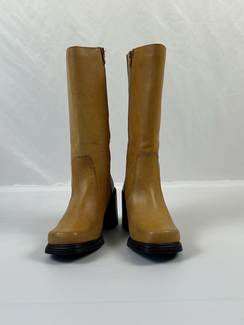 Puede incluir: Un par de botas altas de cuero color canela con puntera cuadrada y tacones de bloque negros. Las botas tienen cierre de cremallera en el lateral. Las botas se exhiben sobre una superficie blanca.