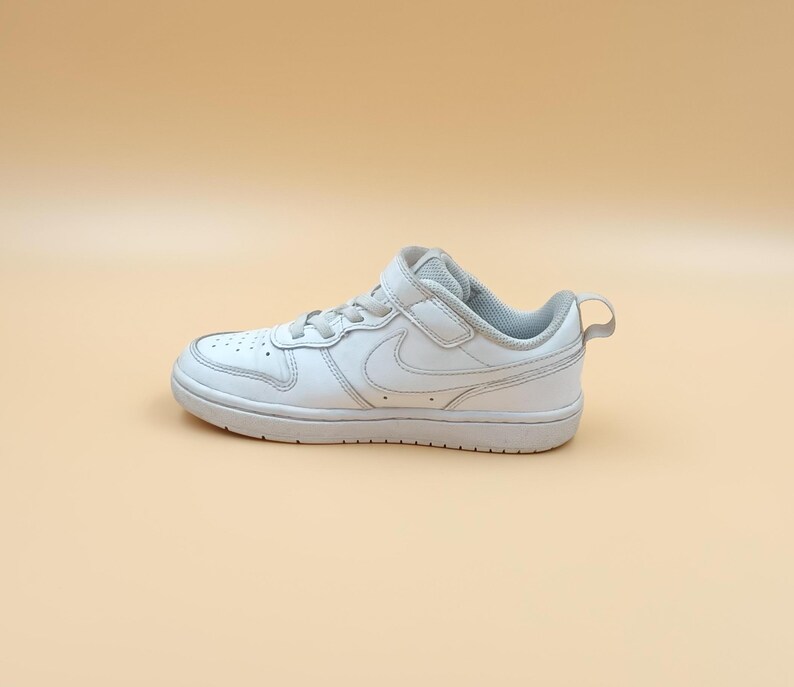 以下が含まれることがあります： ベルクロストラップとナイキのスウッシュロゴが付いた白い革製のローカットスニーカー