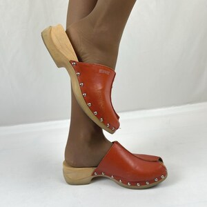 May include: A pair of orange leather clogs featuring a wooden sole and silver-toned metal studs along the base. The upper leather section includes a small embossed Esprit logo. These slip-on shoes have an open-toe design and a low wooden heel.