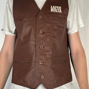 May include: A brown leather vest with a V-neck and four wooden buttons. The vest has two front pockets and the text "LAPIN KULTA" printed on the upper right side.