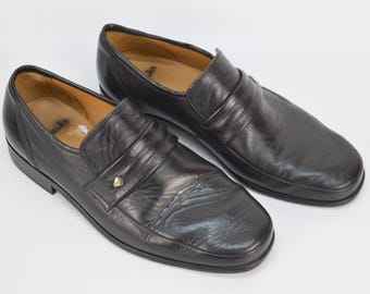Puede incluir: Un par de zapatos de vestir negros de cuero con un acento dorado en la parte delantera. Los zapatos tienen un diseño sencillo y una punta redonda.