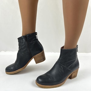 Puede incluir: Un par de botines de cuero negro con punta redondeada, cierre de cremallera lateral y tacón de bloque de madera apilada. Los botines tienen un acabado texturizado en la parte superior de cuero y una suela de goma marrón claro.