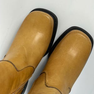 Puede incluir: Un par de botas de cuero color canela con punteras cuadradas y suelas negras. Las botas tienen un aspecto liso y ligeramente desgastado. Las botas son de un color neutro, adecuadas para varios atuendos.