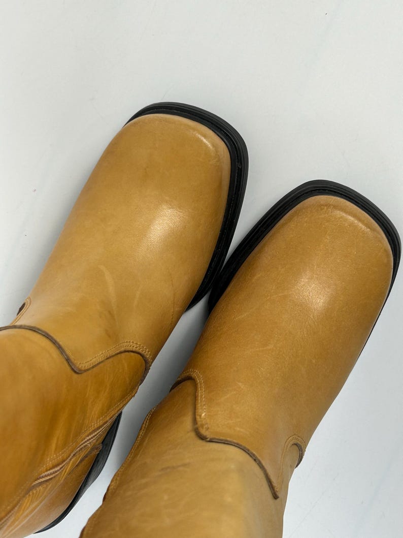 Puede incluir: Un par de botas de cuero color canela con punteras cuadradas y suelas negras. Las botas tienen un aspecto liso y ligeramente desgastado. Las botas son de un color neutro, adecuadas para varios atuendos.