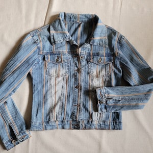 Puede incluir: Una chaqueta vaquera azul con rayas blancas y marrones. La chaqueta tiene cierre de botones y dos bolsillos delanteros.
