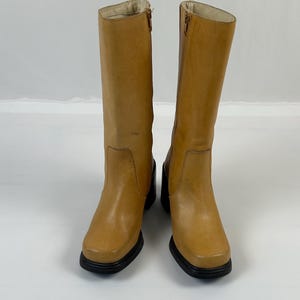 Puede incluir: Un par de botas altas de cuero color canela con puntera cuadrada y suelas negras. Las botas tienen una cremallera lateral y un tac&oacute;n peque&ntilde;o. Las botas est&aacute;n sobre un fondo blanco.