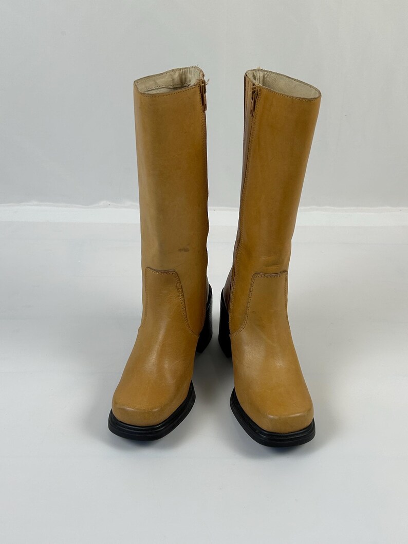 Puede incluir: Un par de botas altas de cuero color canela con puntera cuadrada y suelas negras. Las botas tienen una cremallera lateral y un tac&oacute;n peque&ntilde;o. Las botas est&aacute;n sobre un fondo blanco.