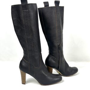 May include: A pair of black leather knee-high boots with a textured finish. The boots have a side zipper, a wooden heel, and a pull tab at the top. The heel height is approximately 3 inches.