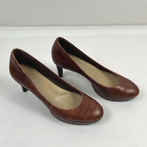 May include: A pair of brown pumps with a textured crocodile-embossed finish, featuring a rounded toe, a low heel, and a beige interior insole with perforated detailing and the text ADIPRENE by adidas printed on the heel area of the shoe.