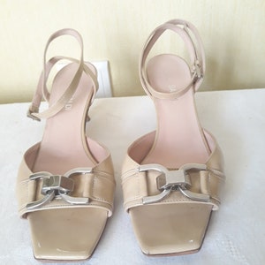 May include: A pair of beige leather heeled sandals with a silver buckle detail. The sandals have a square toe and an ankle strap.