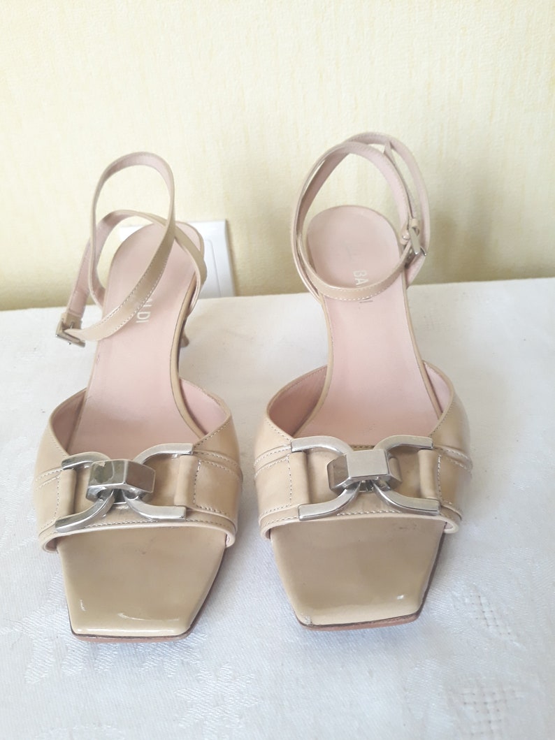 May include: A pair of beige leather heeled sandals with a silver buckle detail. The sandals have a square toe and an ankle strap.