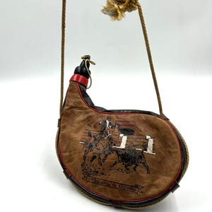 Puede incluir: Cantimplora de cuero marrón con ribete negro y rojo, con una escena de corrida. Tiene una correa de cuerda y una borla decorativa. El texto "Recuerdo de España" es visible en la parte inferior.