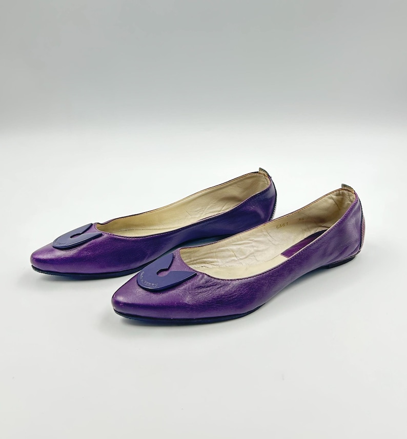 Op de afbeelding: Een paar paarse leren ballerina's met een spitse neus en een donkerpaarse accent op de wreef. De schoenen worden op een witte achtergrond gepresenteerd.