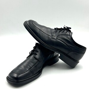 K&ouml;nnte beinhalten: Ein Paar schwarze Lederschuhe mit eckiger Spitze und Ziern&auml;hten. Die Schuhe haben Schn&uuml;rsenkel und einen niedrigen Absatz, geeignet f&uuml;r formelle Anl&auml;sse. Sie werden vor wei&szlig;em Hintergrund pr&auml;sentiert.
