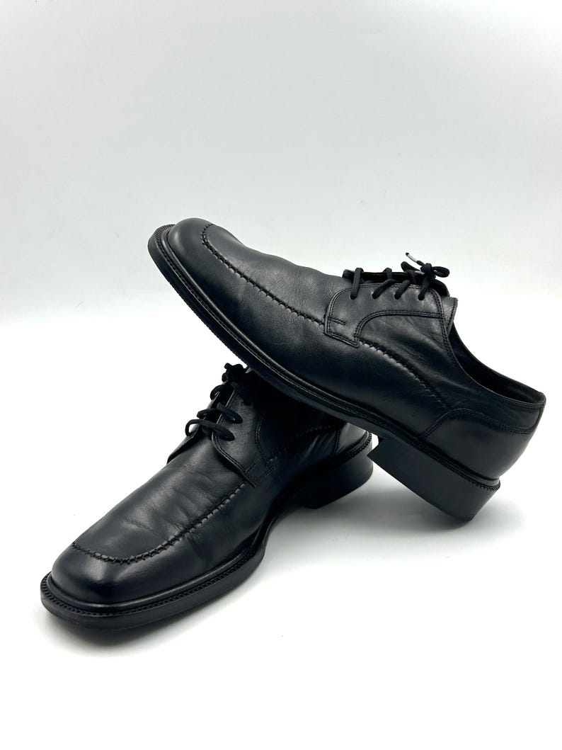 K&ouml;nnte beinhalten: Ein Paar schwarze Lederschuhe mit eckiger Spitze und Ziern&auml;hten. Die Schuhe haben Schn&uuml;rsenkel und einen niedrigen Absatz, geeignet f&uuml;r formelle Anl&auml;sse. Sie werden vor wei&szlig;em Hintergrund pr&auml;sentiert.