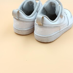 以下が含まれることがあります： 側面にナイキのロゴが付いた、ベルクロ留めの白い革のスニーカーのペアです。