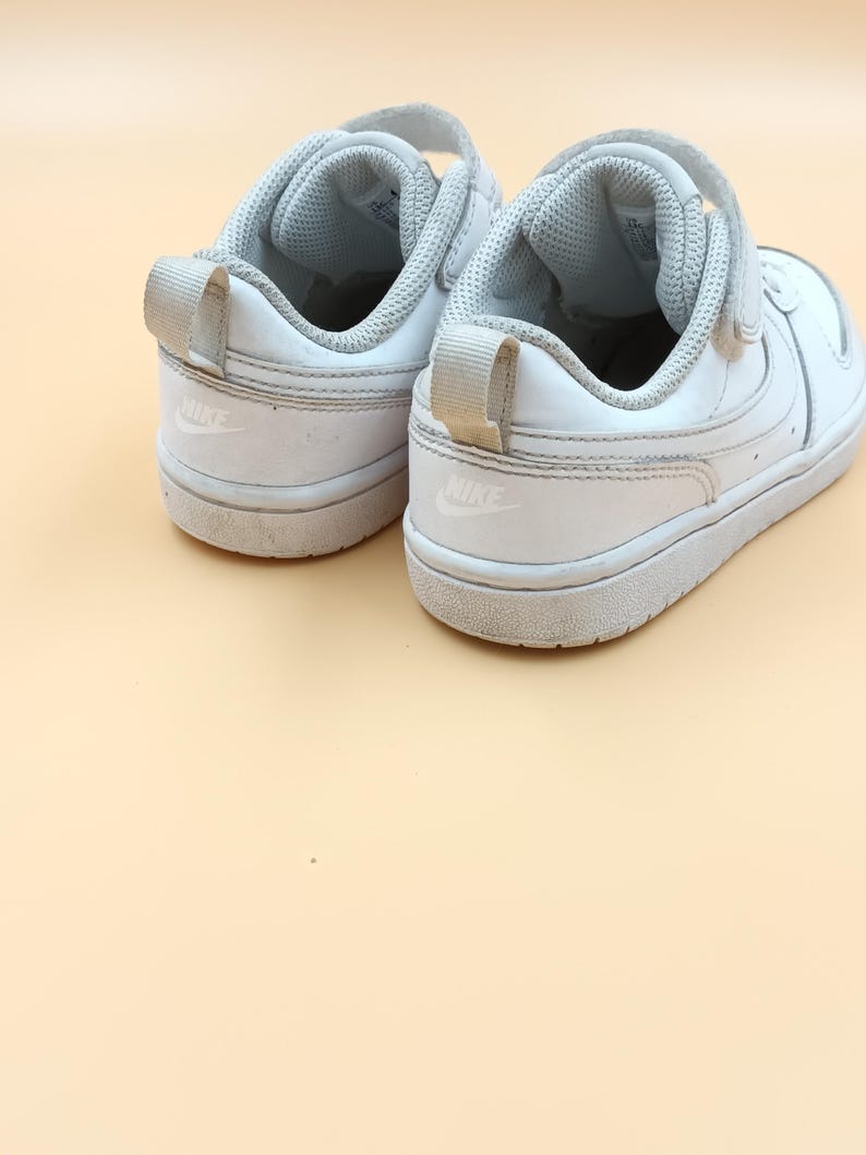 以下が含まれることがあります： 側面にナイキのロゴが付いた、ベルクロ留めの白い革のスニーカーのペアです。