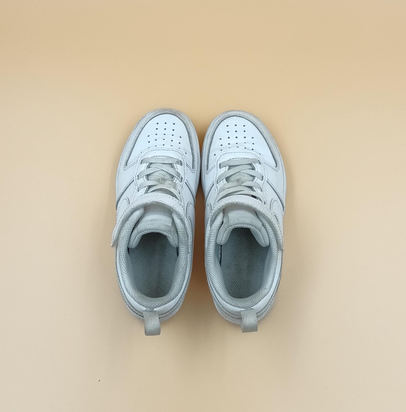 以下が含まれることがあります： ベルクロ留めの白い革のスニーカーのペア。スニーカーは白いゴム製のソールで、薄い茶色の背景の上にあります。