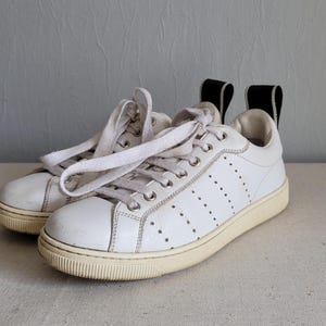 May include: A pair of white leather sneakers with white laces. The shoes have a perforated design on the sides and a cream-colored sole. Black pull tabs are on the back of the shoes.