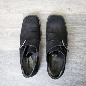 K&ouml;nnte beinhalten: Ein Paar schwarze Wildlederschuhe mit Schnalle und quadratischer Spitze. Die Schuhe haben eine silberne Schnalle und stehen auf einem hellen Holzboden.