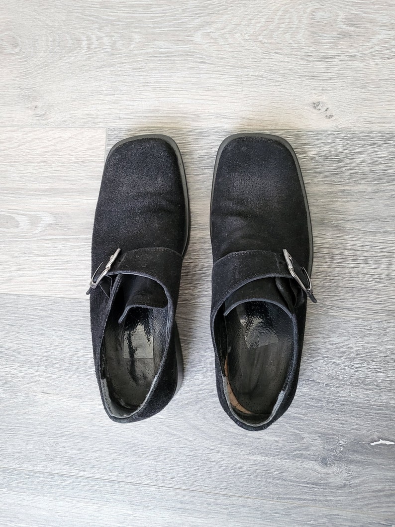 K&ouml;nnte beinhalten: Ein Paar schwarze Wildlederschuhe mit Schnalle und quadratischer Spitze. Die Schuhe haben eine silberne Schnalle und stehen auf einem hellen Holzboden.