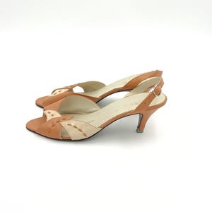 Puede incluir: Un par de zapatos de tacón slingback vintage con punta abierta. Los zapatos presentan una parte superior de cuero marrón con detalles en crema y un tacón pequeño. El diseño incluye recortes decorativos en la correa del dedo del pie y un cierre de hebilla.