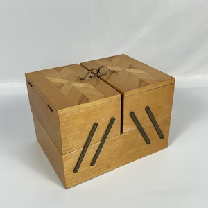 May include: A light-colored wooden sewing box with a hinged lid and brass-colored hardware. The lid features a decorative inlaid pattern. The box is divided into two compartments and has a handle for carrying.