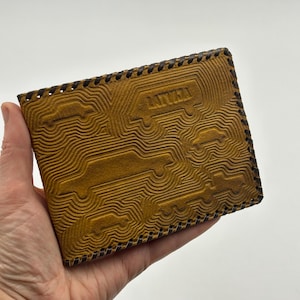 May include: A tan leather wallet with a textured design featuring cars and the word "LATVIA". The wallet has a dark brown stitched border and is held in a hand.