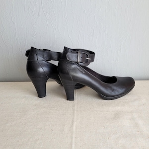 Könnte beinhalten: Ein Paar schwarze Lederschuhe mit Absatz, Knöchelriemen und Schnallen. Die Schuhe haben eine geschlossene Spitze und einen niedrigen Absatz.