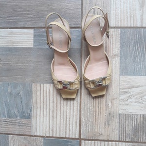 May include: A pair of yellow leather heeled sandals with a buckle detail. The sandals have the brand name "BELLA BALDI" written on the straps.