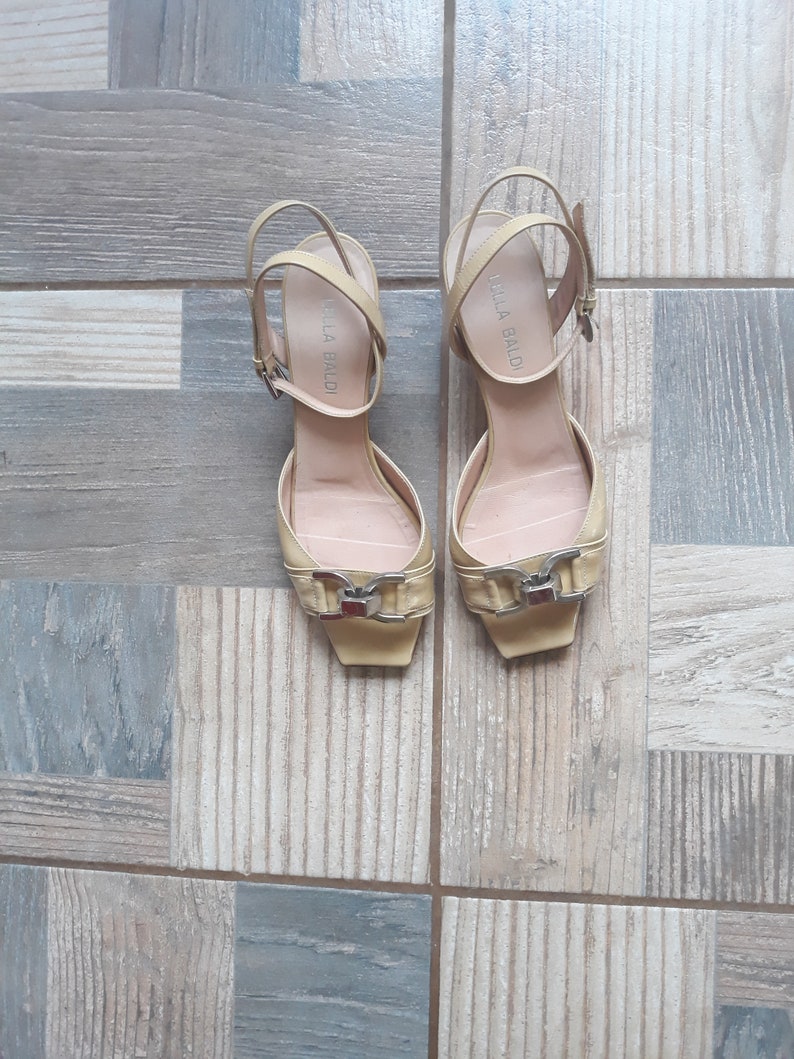 May include: A pair of yellow leather heeled sandals with a buckle detail. The sandals have the brand name "BELLA BALDI" written on the straps.