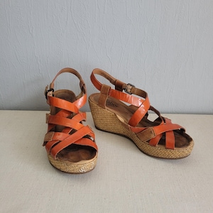 May include: A pair of orange leather wedge sandals with a woven platform. The sandals have multiple straps across the foot and ankle.