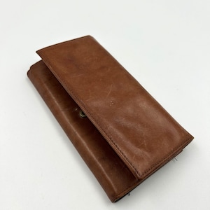 Puede incluir: Una cartera de cuero marrón con cierre de botón a presión. La cartera rectangular tiene un aspecto ligeramente desgastado. El cuero tiene un tono rico y cálido, y las costuras son visibles en los bordes. La cartera está cerrada.