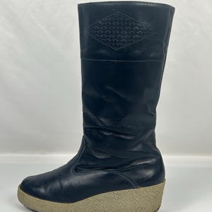 May include: Black leather knee-high boot with a beige wedge sole. The boot features a diamond-shaped woven design at the top. The sole is approximately 2 inches high.