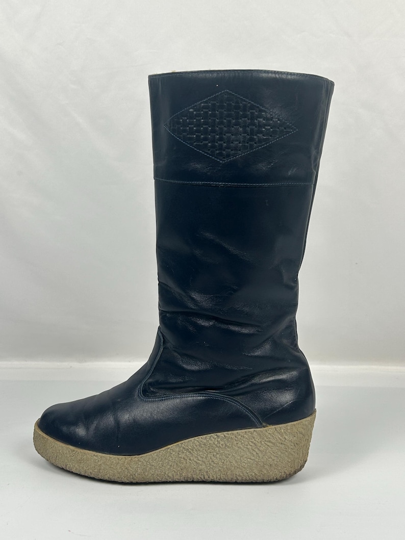 May include: Black leather knee-high boot with a beige wedge sole. The boot features a diamond-shaped woven design at the top. The sole is approximately 2 inches high.