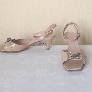 May include: A pair of beige heeled sandals with a silver buckle detail. The sandals have a peep toe and an ankle strap.