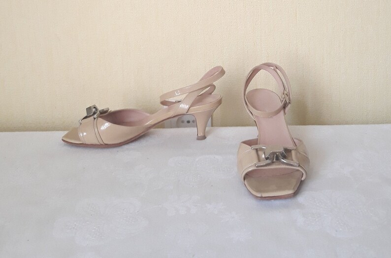 May include: A pair of beige heeled sandals with a silver buckle detail. The sandals have a peep toe and an ankle strap.