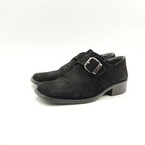 K&ouml;nnte beinhalten: Schwarze Wildlederschuhe mit Schnallenverschluss und silberner Schnalle. Die Schuhe haben einen niedrigen Absatz.