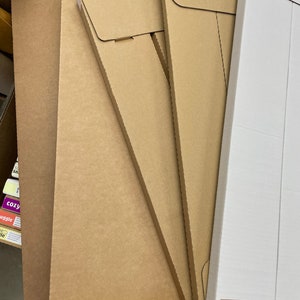 May include: A stack of brown cardboard envelopes and flat sheets. The envelopes have a flap closure. A white cardboard box is partially visible on the right side of the image. The items are likely for packaging or shipping.