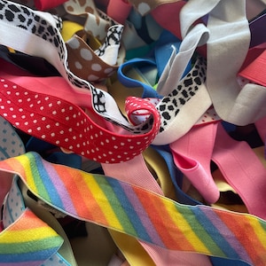 Puede incluir: Primer plano de un montón de cintas elásticas de colores. Las cintas son de varios colores, incluyendo rojo, rosa, amarillo, azul y verde. Algunas cintas tienen lunares o estampados de animales. También se ve una cinta a rayas de arcoíris.