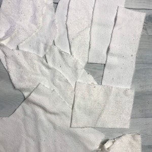 May include: A collection of white fabric pieces, possibly from a towel or similar material, are arranged on a light gray wooden surface. The pieces vary in shape and size, with some appearing to be cut or torn.