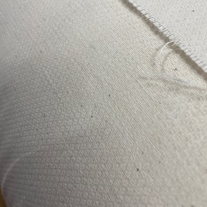 Puede incluir: Primer plano de una tela blanca con un sutil patr&oacute;n texturizado. La tela tiene un borde sin rematar y una costura que recorre la superficie en diagonal.