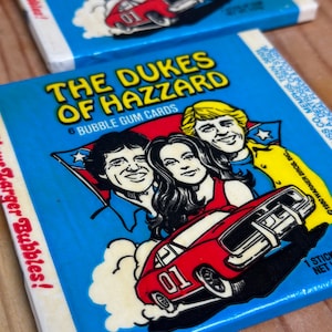 May include: A blue and yellow card with the text "The Dukes of Hazzard" and a picture of a red car with the number "01" on the side. The card also features three people, two men and one woman, with the text "6 Bubble Gum Cards" below the car.