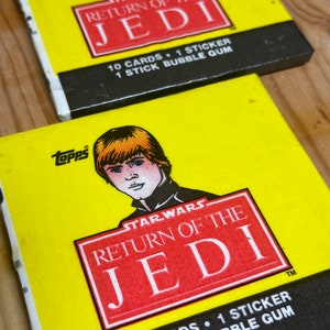 May include: A yellow and black card with a red and white logo that says "Star Wars Return of the Jedi". The card has a picture of a young Luke Skywalker with blonde hair and blue eyes. The text on the card says "10 cards, 1 sticker, 1 stick bubble gum".