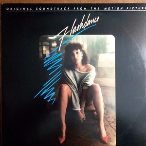 May include: A black vinyl record album cover with the title "Flashdance" in white and blue lettering. The cover features a photo of a woman wearing a grey sweater and red shoes, sitting on a wooden surface.