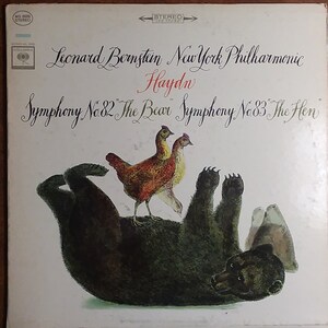 May include: A vintage vinyl record cover featuring a black bear lying on its back with three chickens standing on its belly. The cover is for a recording of Haydn's Symphonies No. 82 "The Bear" and No. 83 "The Hen" performed by Leonard Bernstein and the New York Philharmonic.