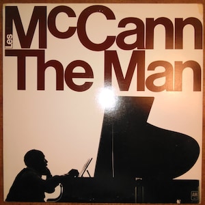 May include: A vintage vinyl record album cover featuring a silhouette of a person playing a grand piano. The album title is "Les McCann The Man" in bold, brown text.