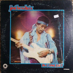 May include: A vintage vinyl record album cover featuring Jimi Hendrix playing a guitar on stage. The album cover is black with a blue border and the text "Jimi Hendrix In Concert" in white letters. The album cover is in stereo.