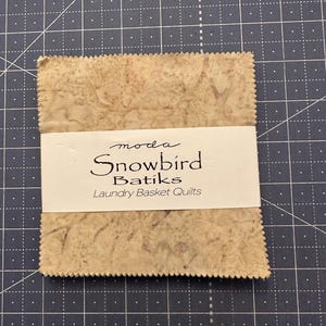 May include: A stack of Snowbird Batiks fabric squares, featuring a mottled beige and brown pattern. The fabric is wrapped with a white label that reads "moda Snowbird Batiks Laundry Basket Quilts."